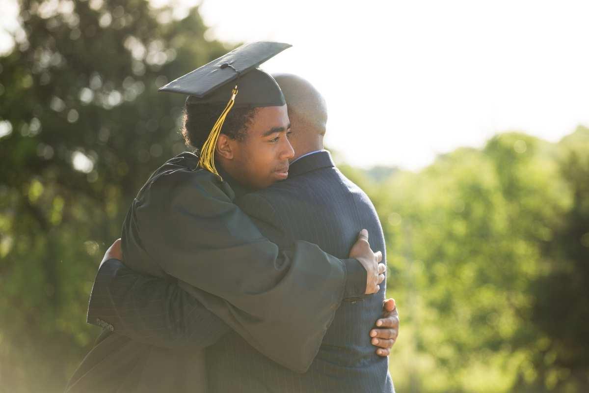 male graduate in cap and gown hugging person